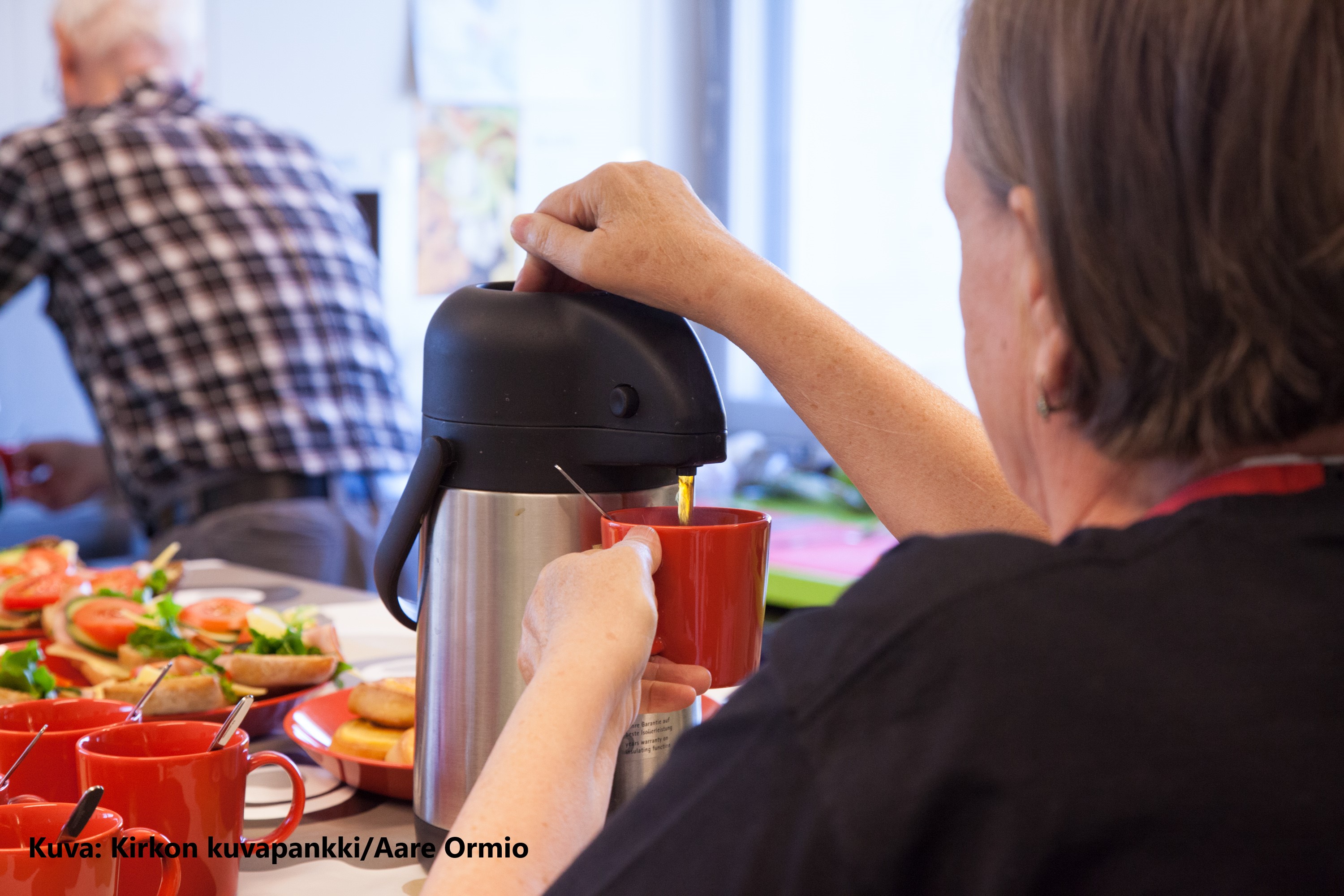 Iäkäs nainen kaataa kahvia termoskannusta kuppiin ja taustalla iäkkään miehen hahmo.