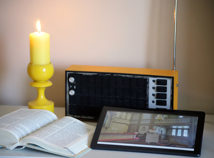 Sanajumalanpalvelus radion ja verkon välityksellä Pöydällä palava kynttilä, avoin Raamattu, radio sekä tablettitietokone, jonka näytöllä näkyy Kajaanin kirkko