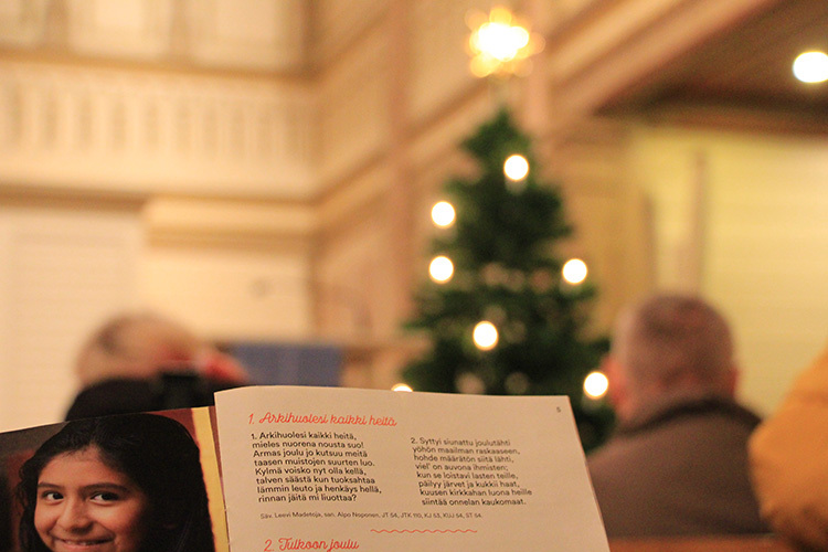 Joululaulutilaisuuden tunnelmaa Kajaanin kirkossa, etualalla lauluvihko, taustalla ihmisiä ja joulukuusi