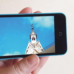 Käsi pitelee matkapuhelinta, jonka näytöllä näkyy Kajaanin kirkon tapuli sekä sininen taivas